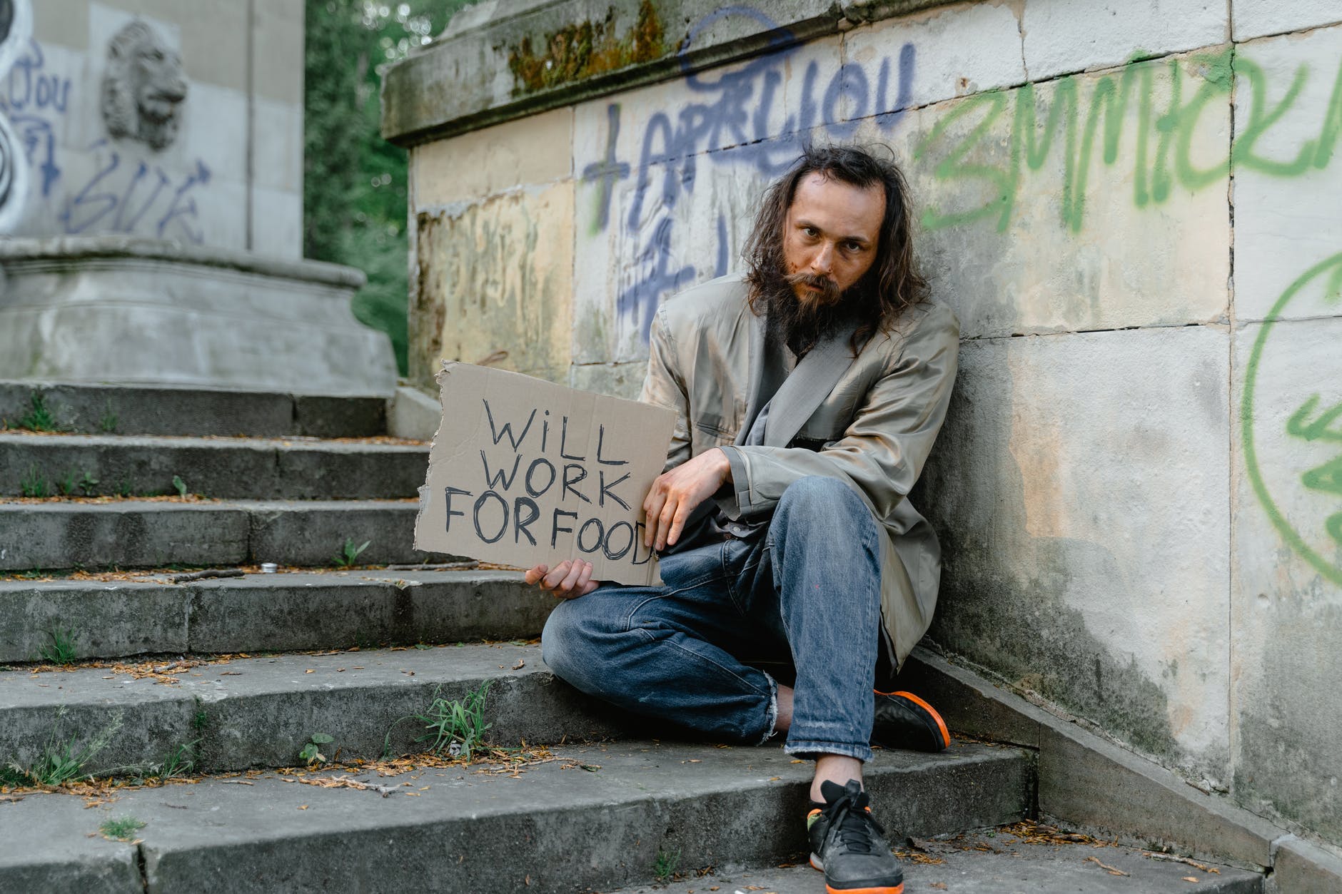 a male beggar sitting on the stairs while holding a will work for food banner