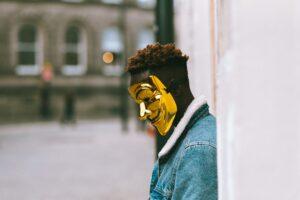 pensive black guy in golden anonymous mask standing on street in daytime